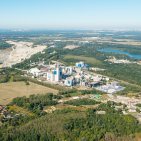 Aerial view of a large industrial plant surrounded by forests and fields, with a lake in the background. Several buildings, silos, and chimneys suggest a cement factory.