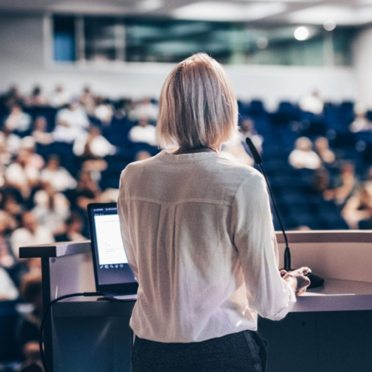 A person standing at a lectern speaking into a microphone to an audience in a conference hall.