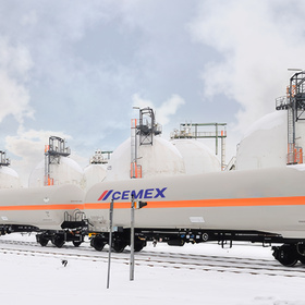 Freight rail cars with white tanks and orange stripes, labeled with a cement company logo, on a snowy industrial site.